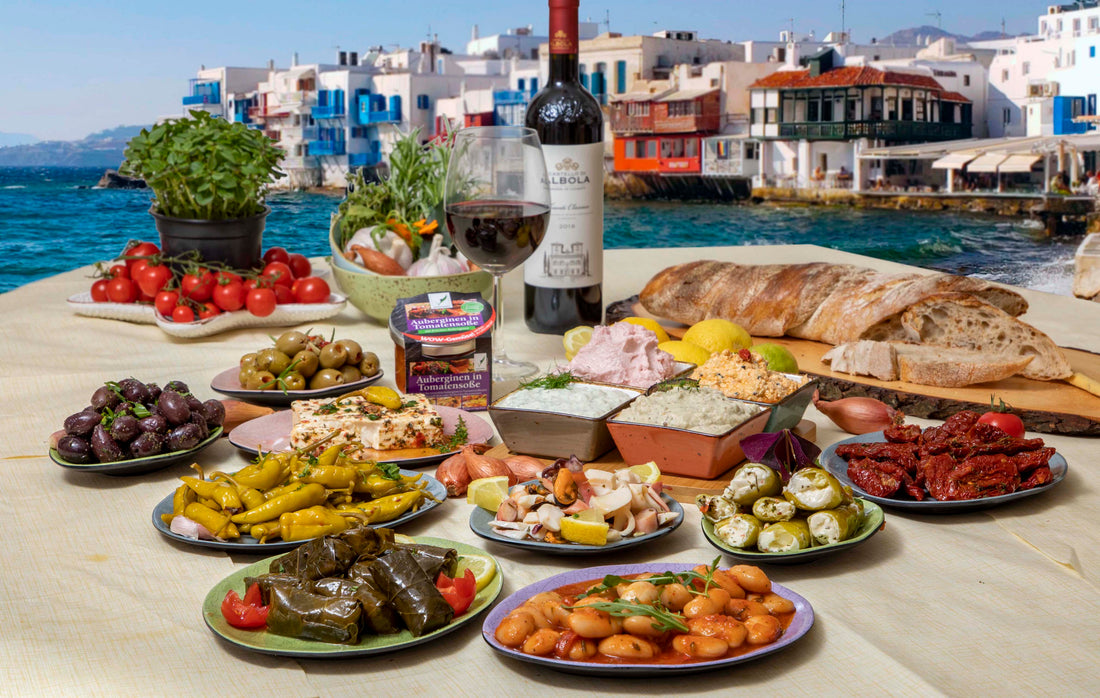 Das Bild zeigt einen Tisch mit etlichen Antipasti und einer Weinflasche. Im Hintergrund sieht man Mykonos.